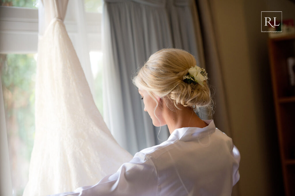 A bride is putting her wedding dress on in front of a window.