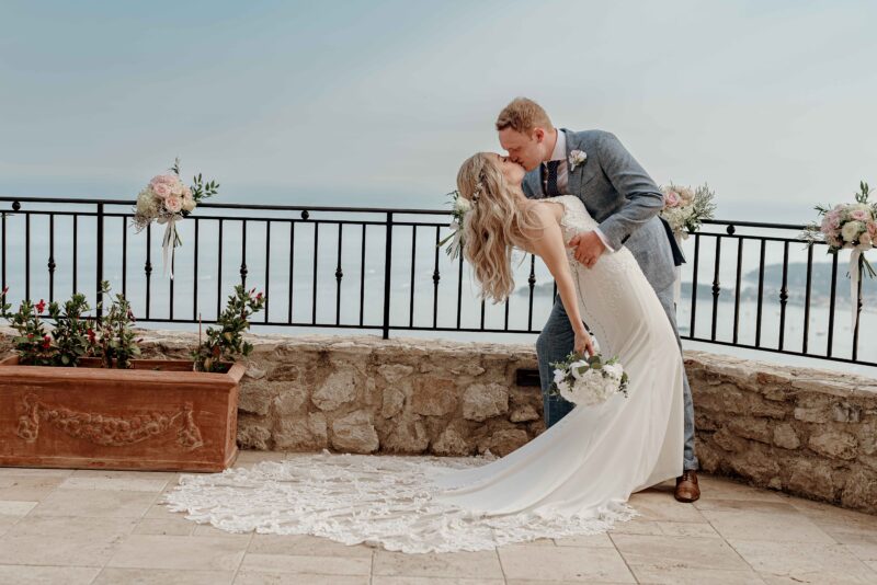 A bride and groom kissing on a balcony overlooking the ocean.