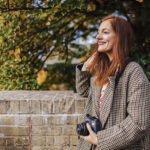 A woman with red hair stands outdoors wearing a checkered coat, holding a camera. She is smiling and standing in front of a brick wall and greenery.