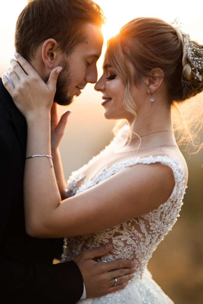 A bride and groom embrace closely at sunset, touching foreheads and smiling, with soft golden light in the background.