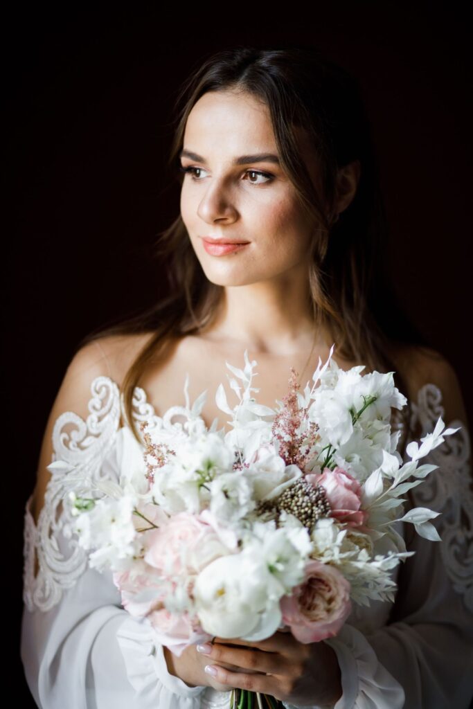 A woman in a white lace wedding dress holds a bouquet of white and pink flowers, looking slightly to the side against a dark background.